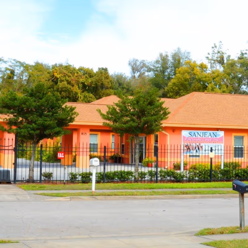 Exterior view of San Jean Facility Care Inc., a single-story building with an orange facade and a brown roof, surrounded by a black metal fence and trees, with a mailbox and a street in the foreground.