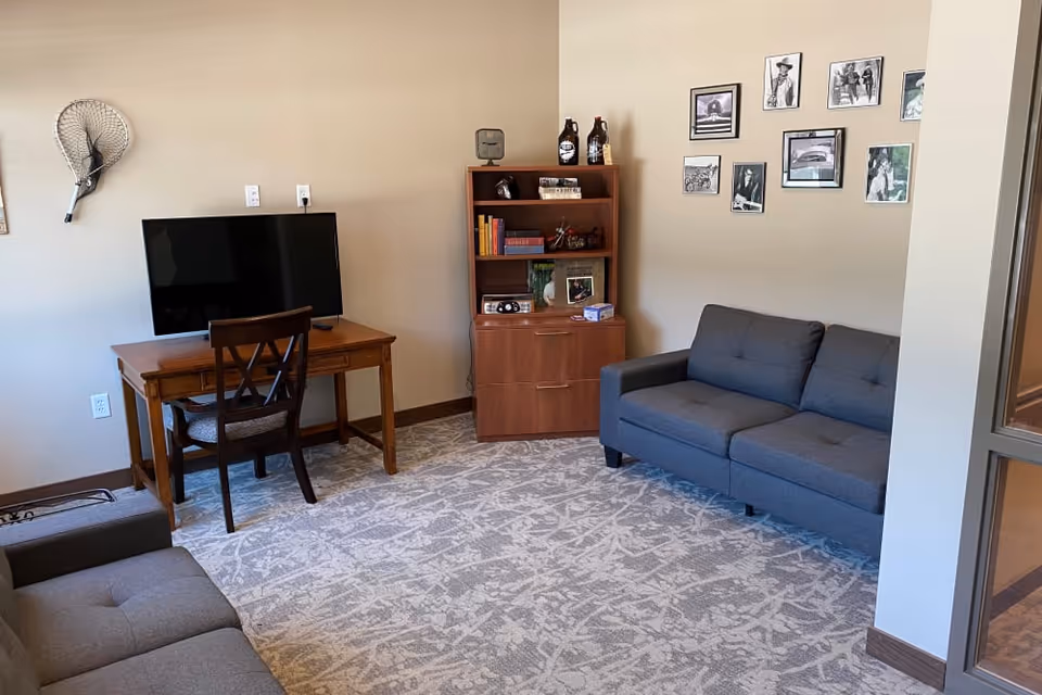 A cozy living room with two gray sofas facing each other, a wooden desk with a chair and a flat-screen TV on it, a wooden bookshelf with books and decorative items, and a wall decorated with several black and white framed photographs.