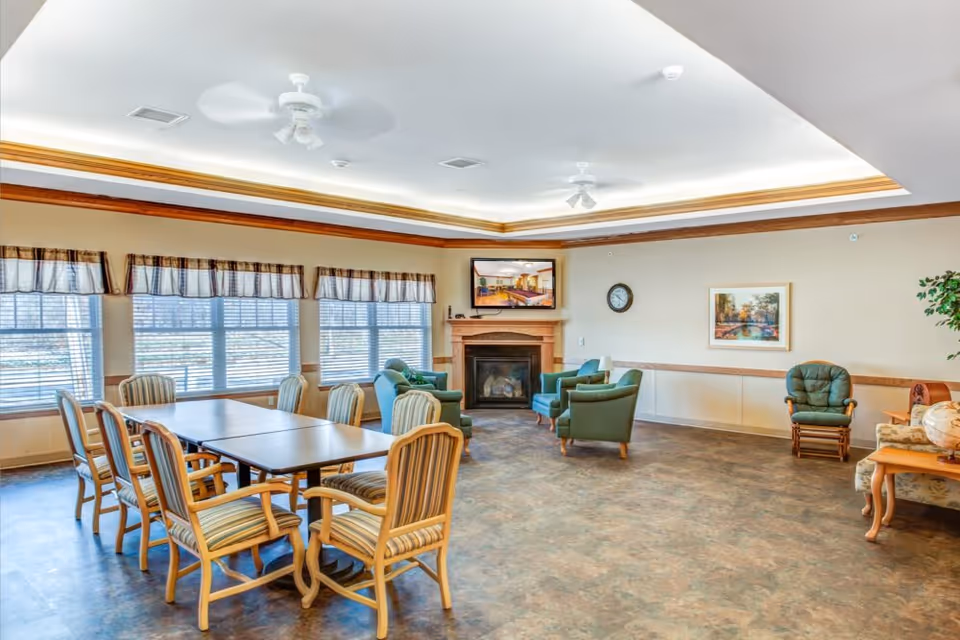 Bright senior living common room with a dining table and chairs, lounge seating around a fireplace, and large windows.