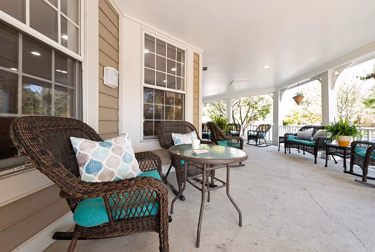 Covered outdoor patio area with wicker chairs and sofas featuring teal cushions and patterned pillows. A round glass-top table is placed between two chairs. The patio has white railings and columns, with hanging plants and greenery visible outside.