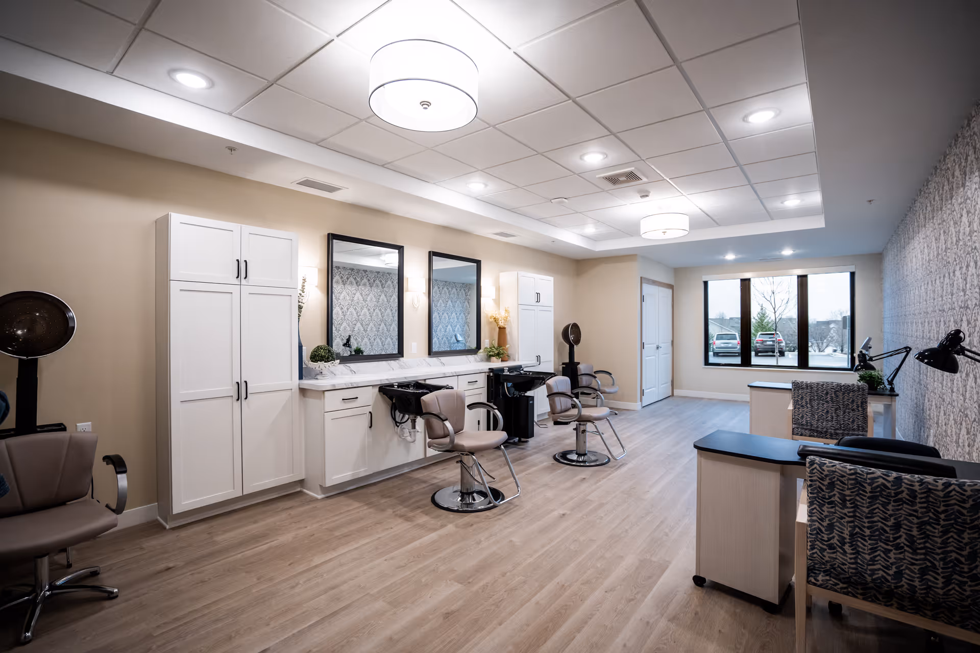 Interior view of a modern salon area in a senior living facility with two styling chairs in front of sinks and mirrors, white cabinetry, a hair dryer, and two desks with chairs near a large window showing parked cars outside.