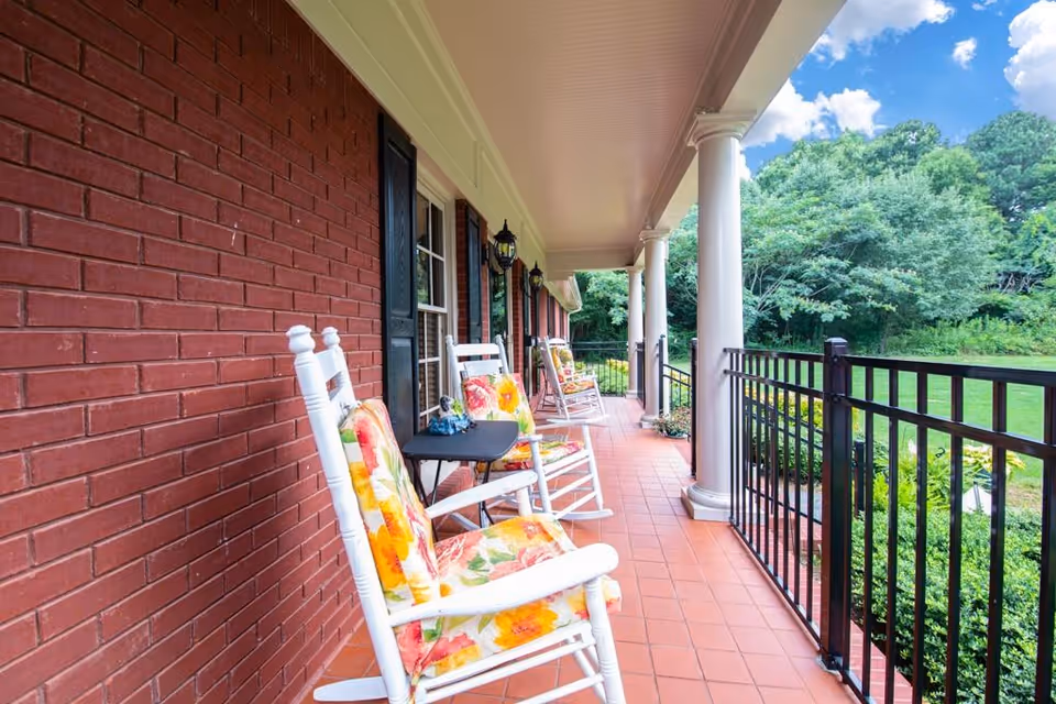 Covered front porch with white rocking chairs with floral cushions, columns, and a black railing overlooking a green lawn.
