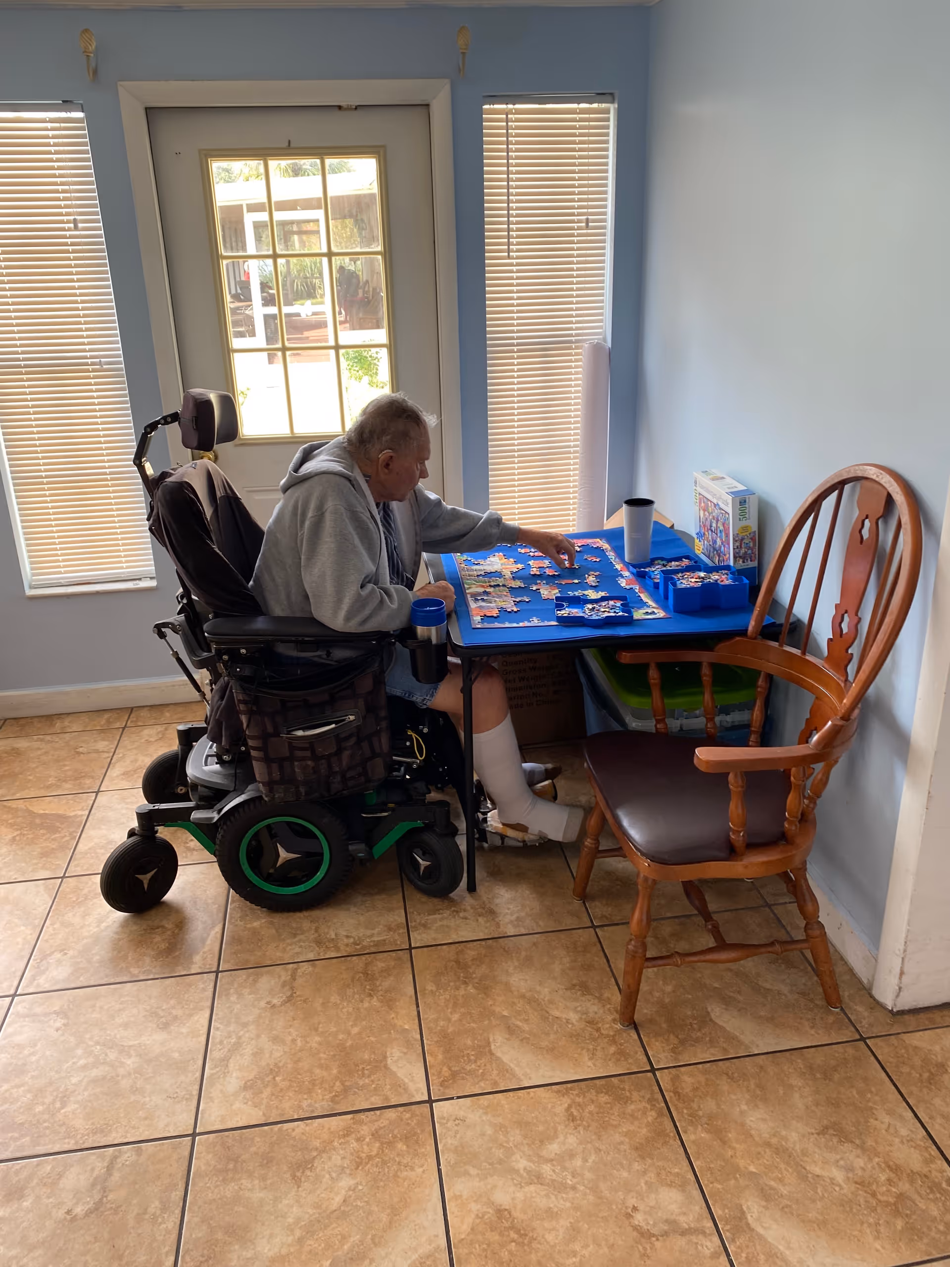 An elderly man in a motorized wheelchair is sitting at a small table working on a jigsaw puzzle. The room has light blue walls, two windows with closed blinds, and a door with glass panes letting in natural light. There is an empty wooden chair next to the table.