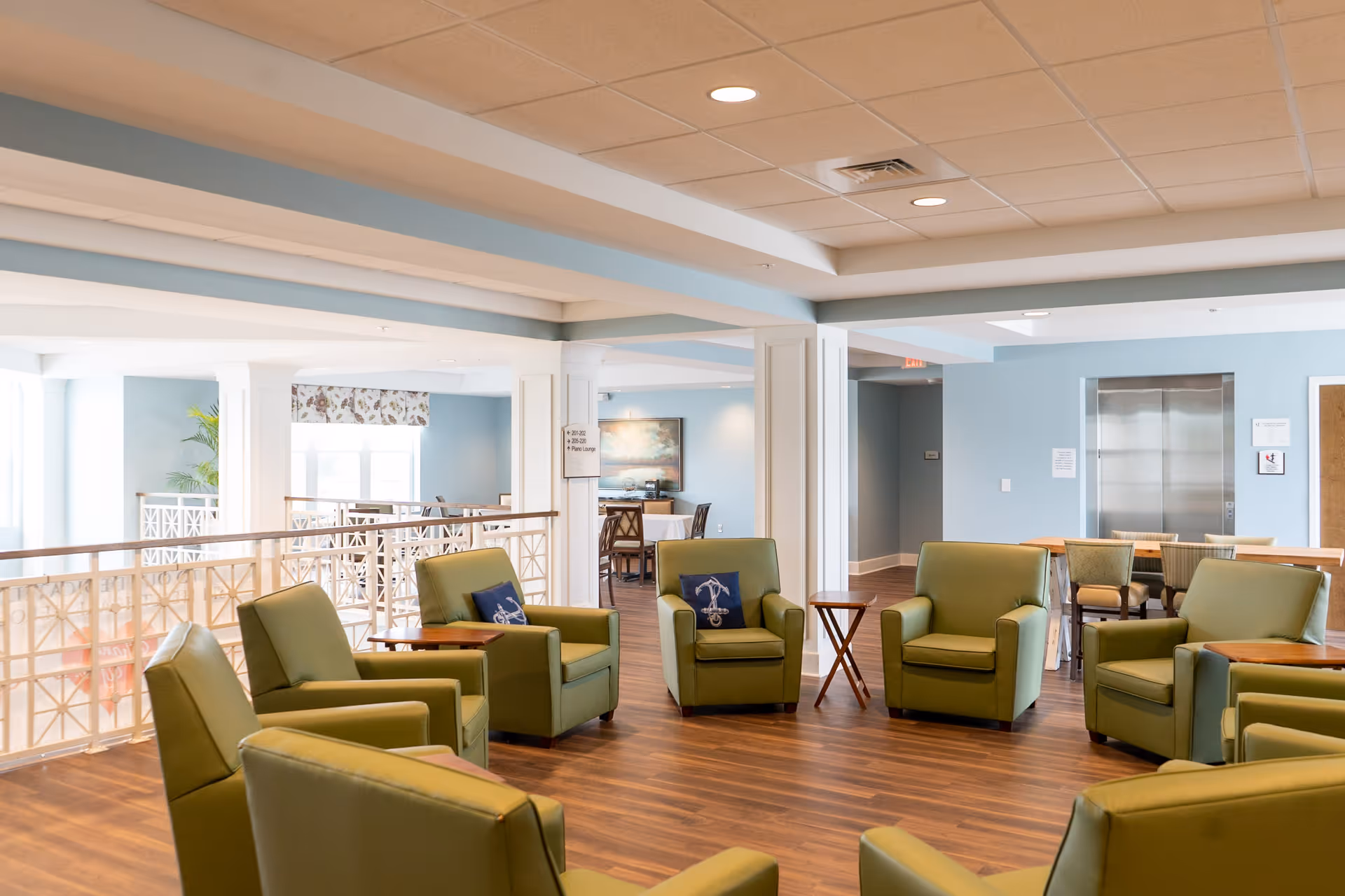 A bright and spacious common area with several green armchairs arranged in a circle on a wooden floor. The room has light blue walls, a white ceiling with recessed lighting, and large windows letting in natural light. There is a small wooden side table between some chairs, and an elevator is visible in the background.