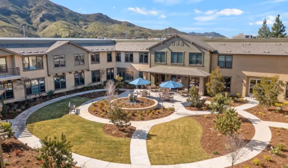 Outdoor courtyard area of Oakmont of Agoura Hills senior living facility with circular walking paths, landscaped garden beds, patio tables with blue umbrellas, and surrounding two-story building with windows. Mountains are visible in the background under a blue sky with some clouds.