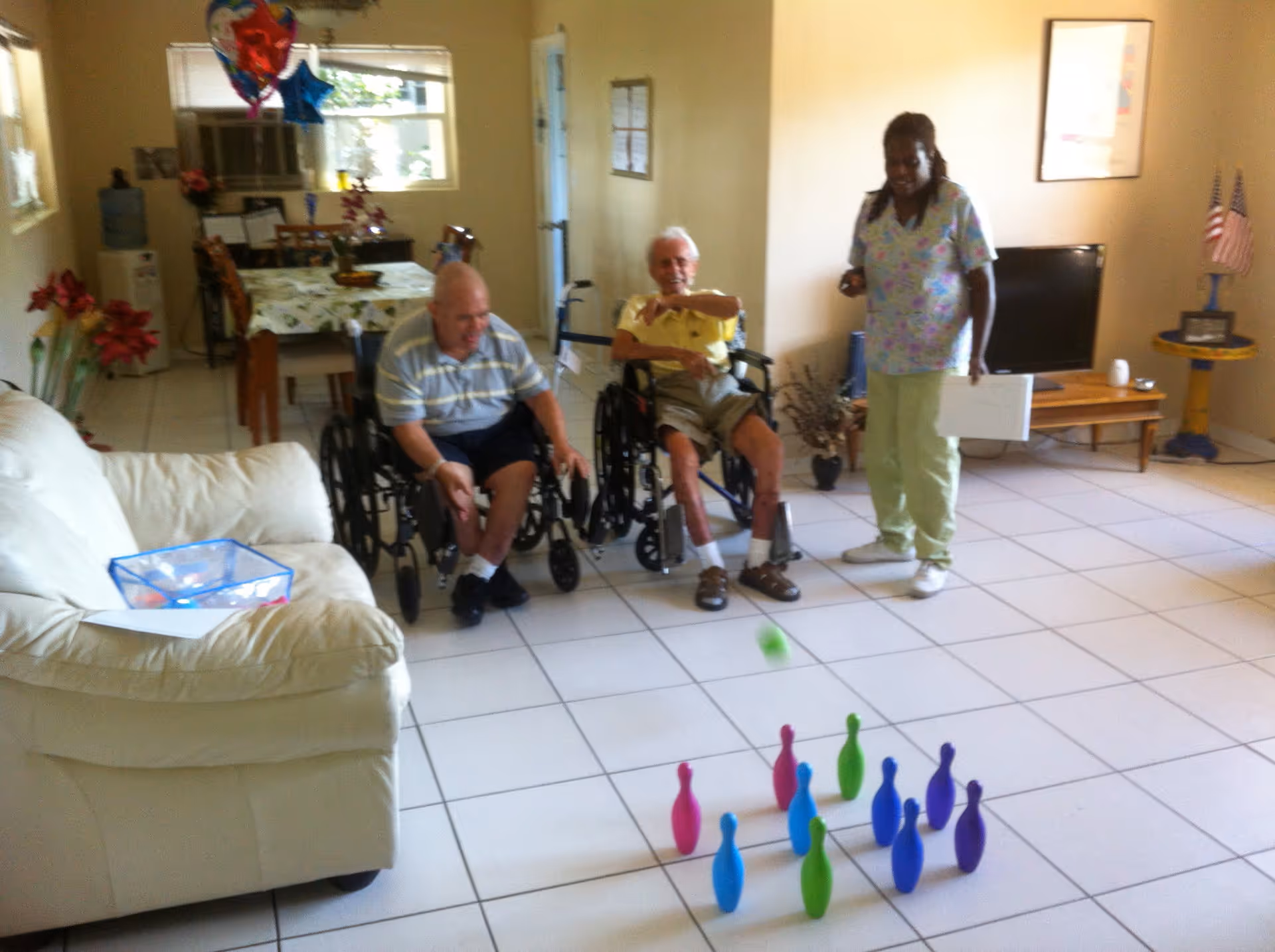 Two elderly men in wheelchairs playing indoor bowling with colorful plastic pins on a tiled floor, assisted by a caregiver standing nearby holding a clipboard in a bright living room with a white couch, dining table, and television.