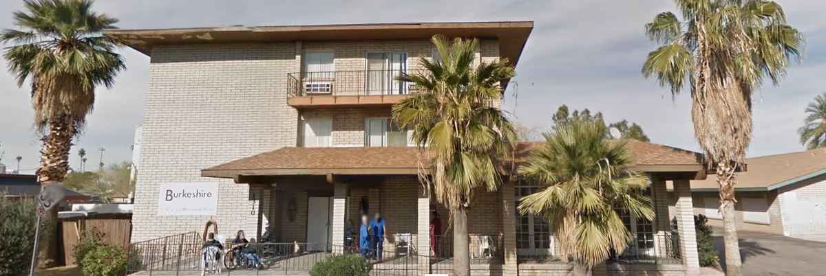 Two-story brick retirement hotel front with a covered entrance, palm trees, a sign reading 'Burkeshire', and people including wheelchair users near the doorway.