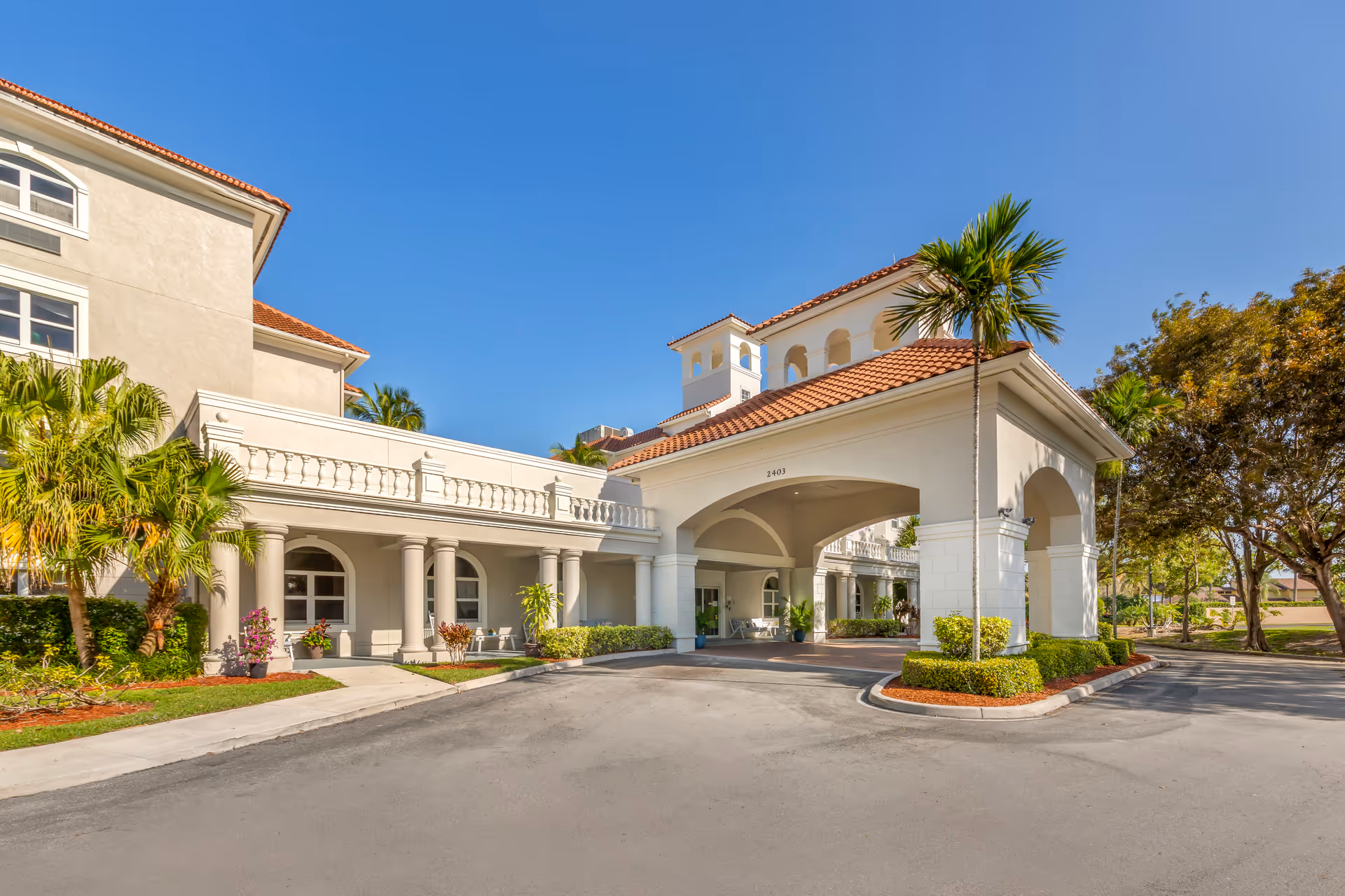 Exterior view of Brookdale Deer Creek facility featuring a large covered entrance with arches, white columns, and a red-tiled roof. Palm trees and landscaped greenery surround the building under a clear blue sky.