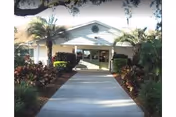 Front exterior view of a single-story building with a covered entrance, surrounded by palm trees and landscaped bushes along a concrete walkway.