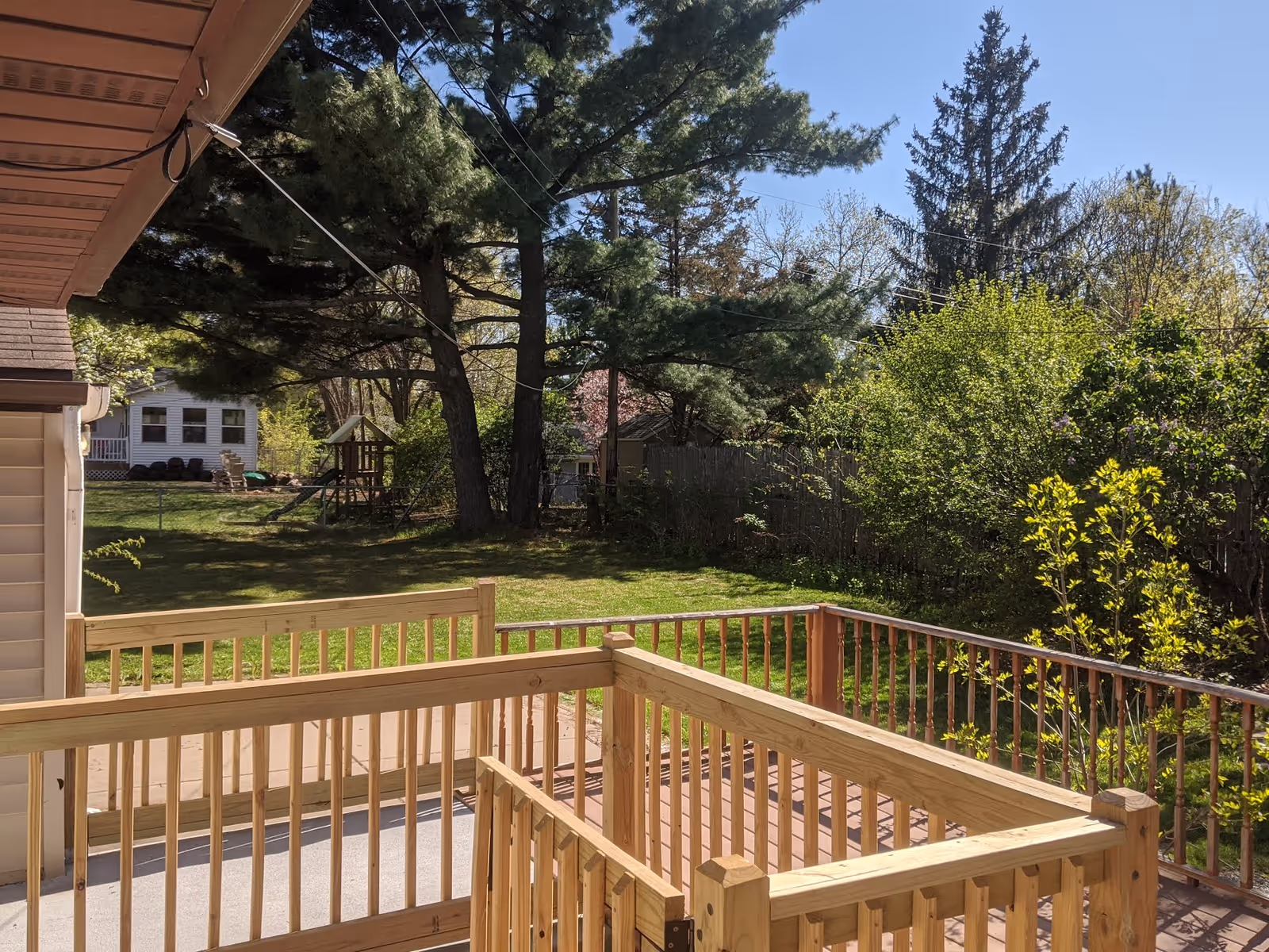View of a wooden deck railing overlooking a green backyard with trees, bushes, and a small play structure. A neighboring house is visible in the background under a clear blue sky.