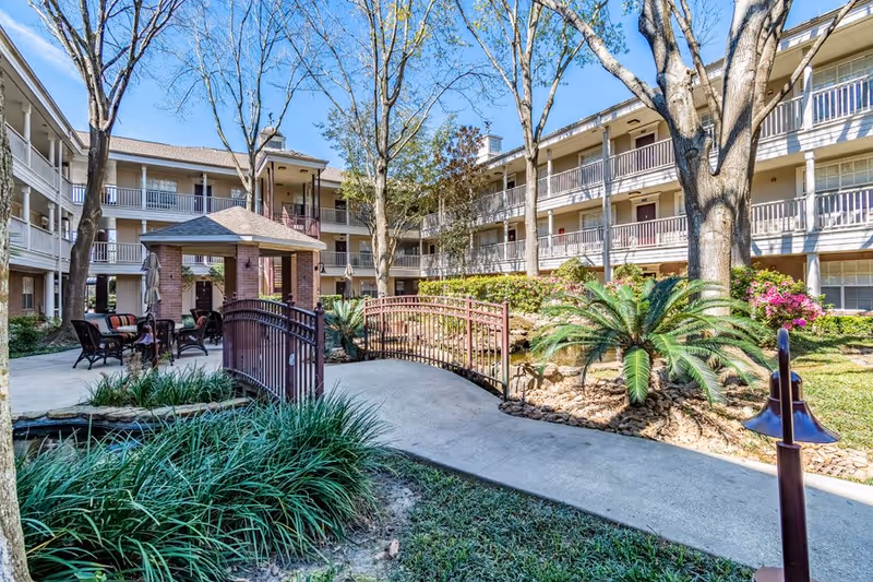 Sunny landscaped courtyard of a three-story senior living building with a small bridge, gazebo, patio seating, and mature trees.