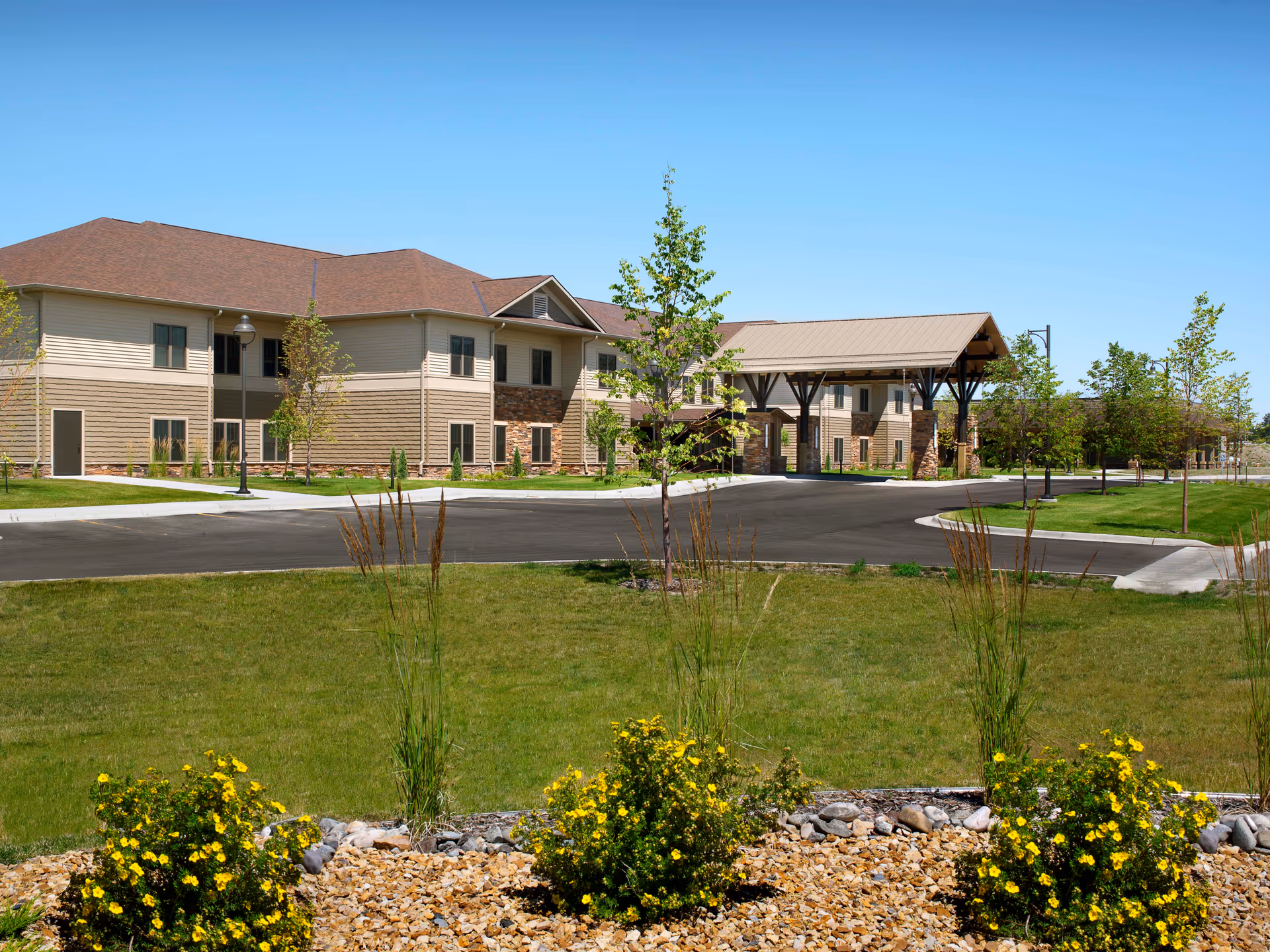 Exterior view of The Grandview at Benefis senior living facility showing a two-story building with beige and brown siding, a covered entrance, a paved driveway, green lawn, small trees, and yellow flowering bushes under a clear blue sky.