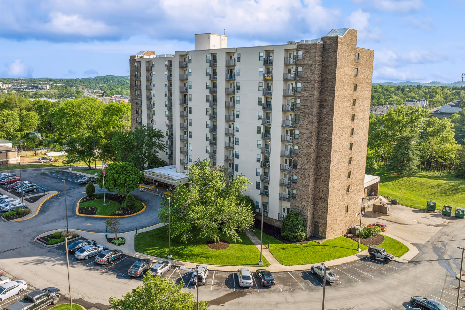 A multi-story senior living facility building named Windlands South surrounded by green trees and a parking lot with several parked cars under a partly cloudy blue sky.