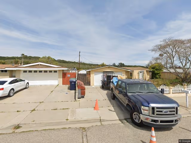 View of a residential driveway with a white car parked on the left and a dark blue pickup truck parked on the right. Two single-car garages are visible, along with a trash bin and recycling bin near the center. There is a small orange traffic cone on the driveway and a white picket fence on the right side. Trees and hills are visible in the background under a partly cloudy sky.