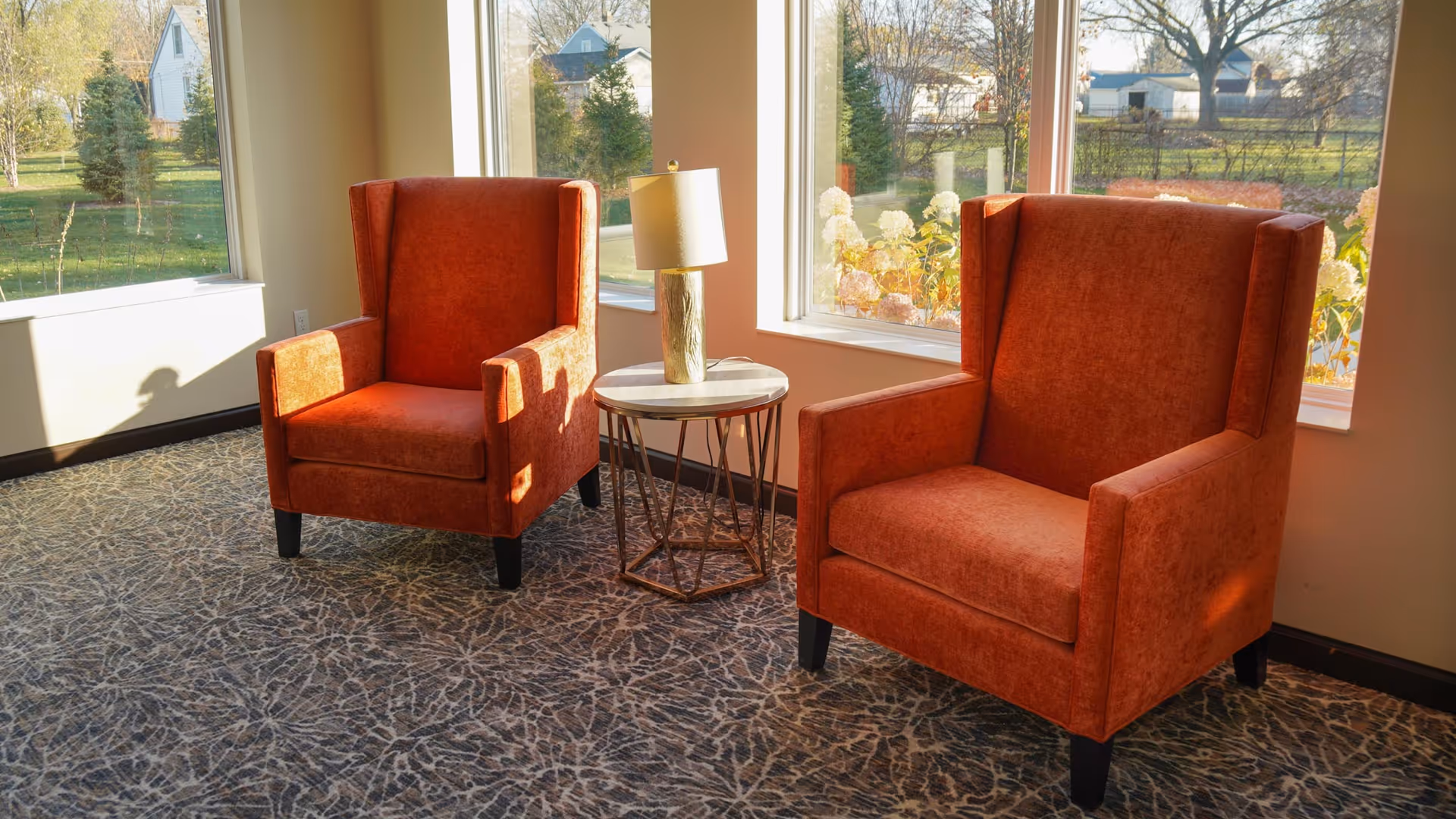 Two orange upholstered armchairs with high backs are positioned near large windows letting in natural light. Between the chairs is a small round side table with a lamp on it. Outside the windows, trees and houses are visible.