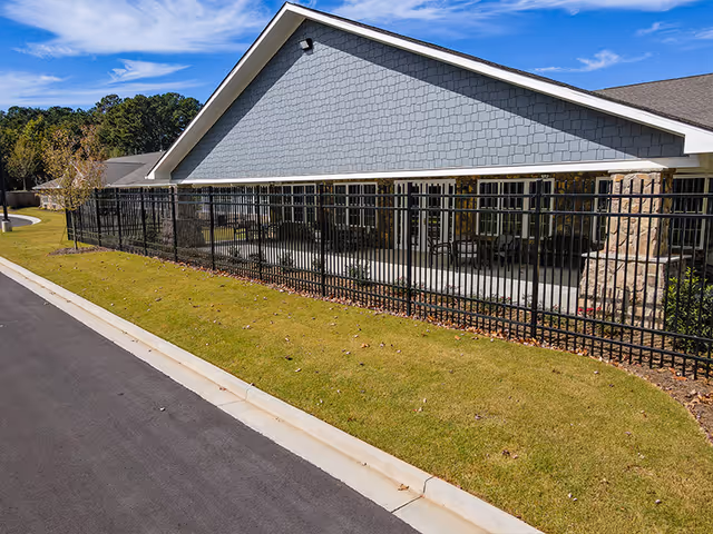 Exterior view of a single-story assisted living building with a fenced patio, lawn, and driveway.