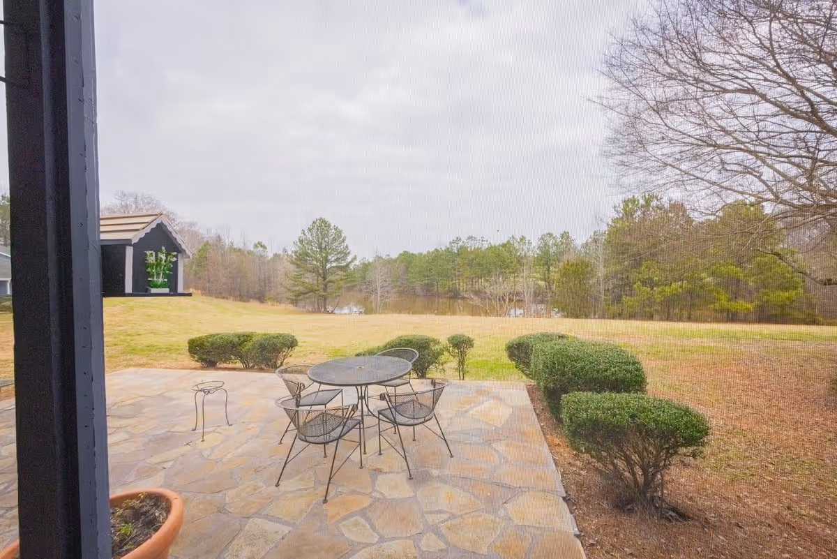 View of an outdoor patio area with a round metal table and four matching chairs on a stone floor. The patio overlooks a grassy yard with neatly trimmed bushes and trees in the background under a cloudy sky.