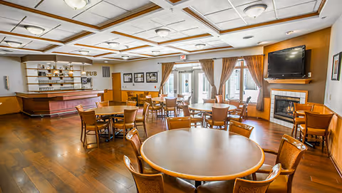 A spacious dining area with multiple round wooden tables and chairs arranged neatly. There is a bar area labeled 'PUB' on the left side with shelves holding glasses and bottles. Large windows with brown curtains allow natural light to fill the room. A flat-screen TV is mounted above a fireplace on the right wall. The ceiling has a grid pattern with recessed lighting fixtures.