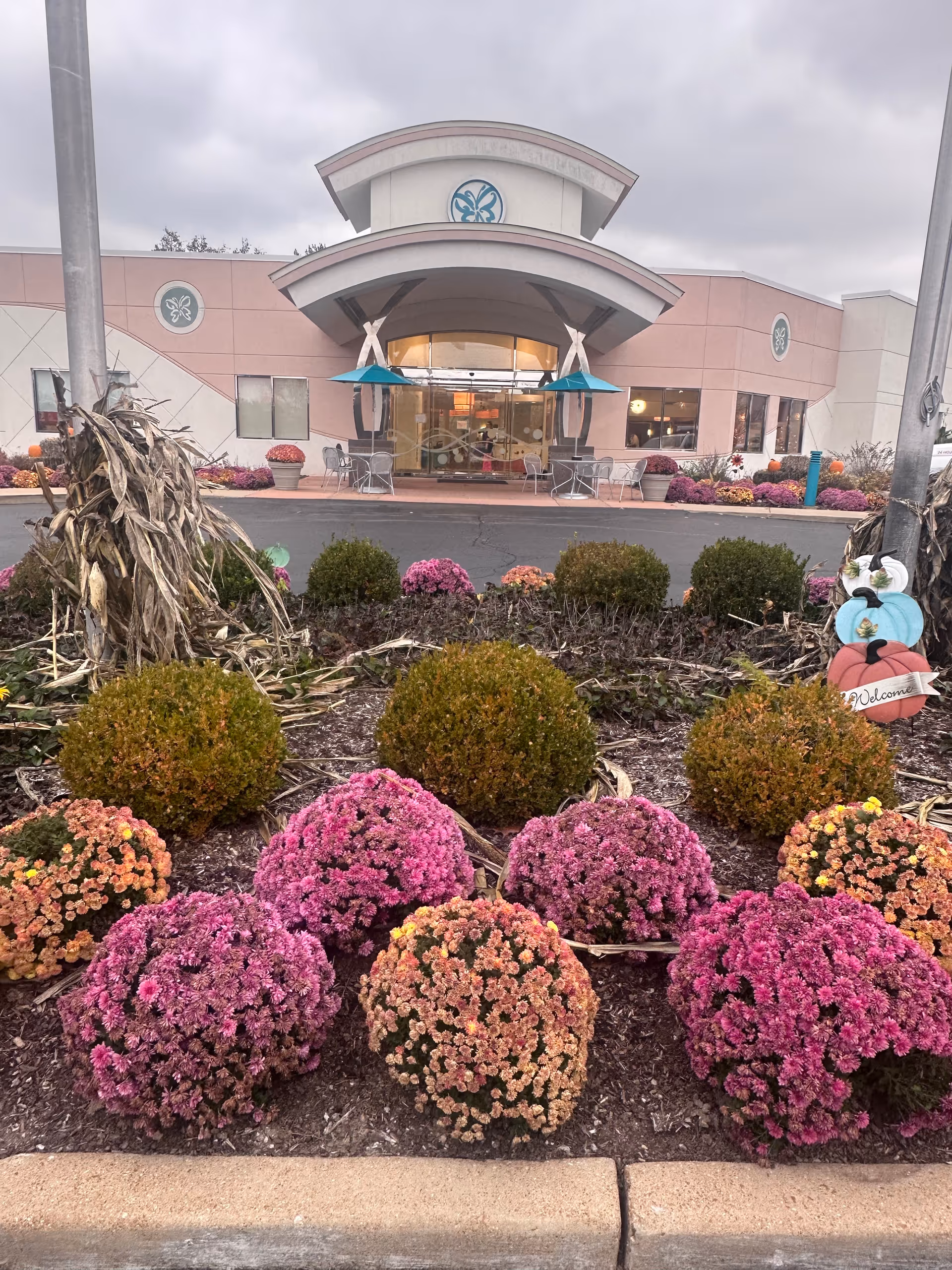 Exterior view of Garden Villas South facility entrance with a landscaped flower bed in the foreground featuring pink and orange flowers, green bushes, and dried corn stalk decorations. The building has a curved roof over the entrance with glass doors and windows, and there are outdoor tables and chairs near the entrance.