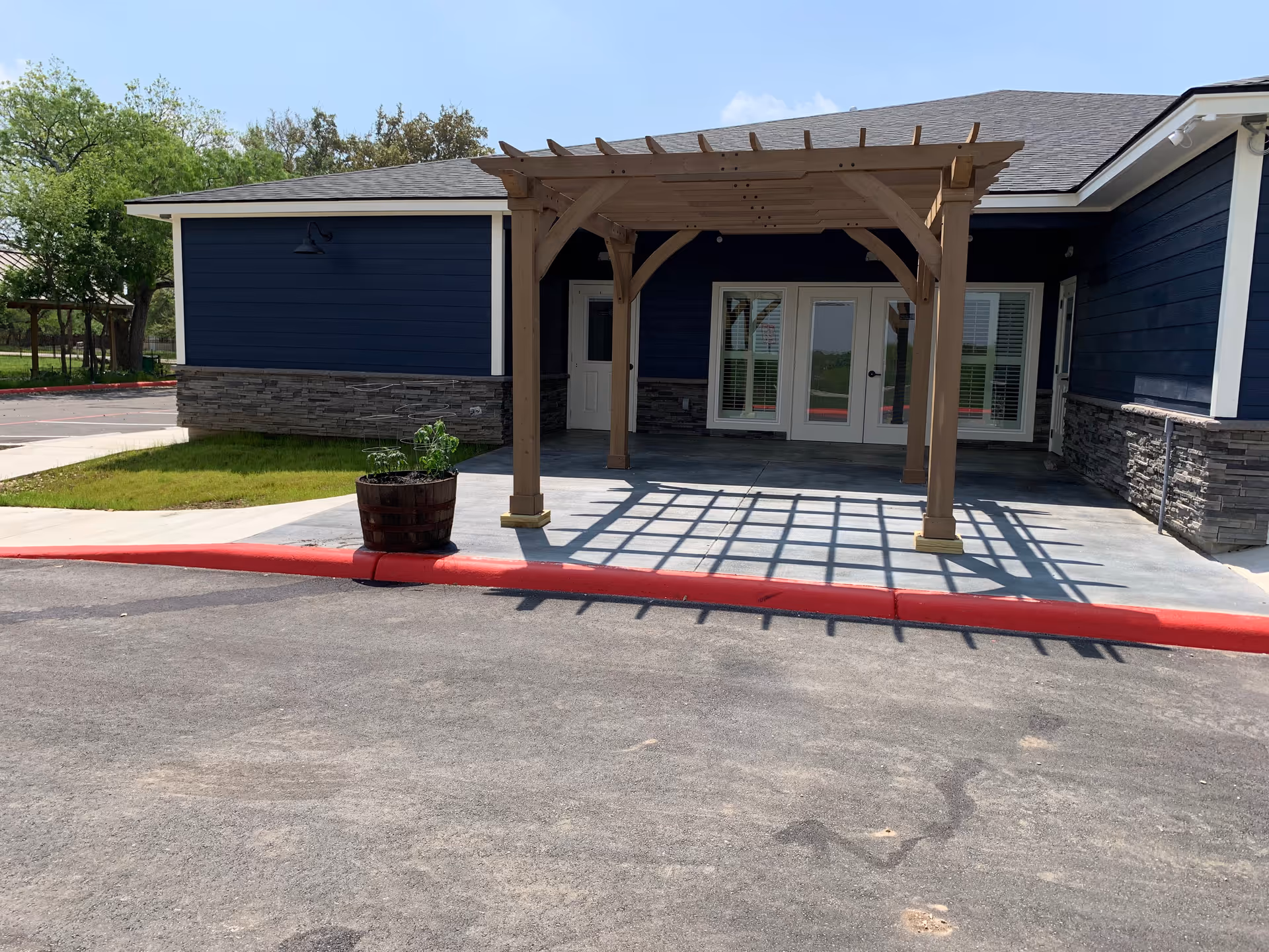 Exterior view of a building with dark blue siding and stone accents, featuring a wooden pergola over a concrete patio area in front of double glass doors. There is a large wooden planter with green plants near the edge of the patio, and a red painted curb along the asphalt driveway. Trees and greenery are visible in the background.
