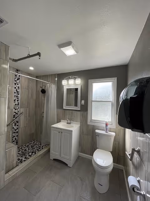 A clean and modern bathroom featuring a walk-in shower with a curtain and mosaic tile floor, a white vanity with a sink and mirror above it, a toilet with a can of air freshener on top, a frosted window letting in natural light, and a wall-mounted black paper towel dispenser.