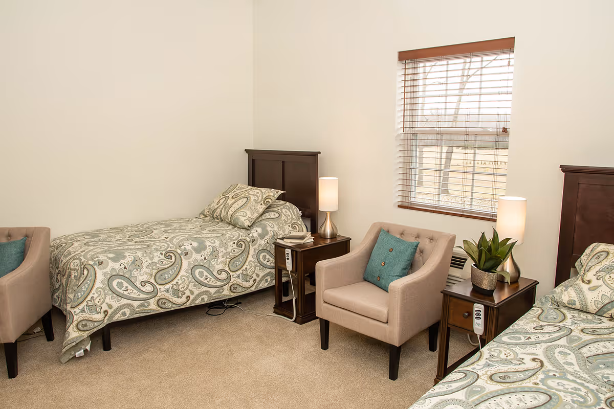 Two twin beds with paisley bedding, upholstered chairs, nightstands with lamps, and a window in a neutral bedroom.