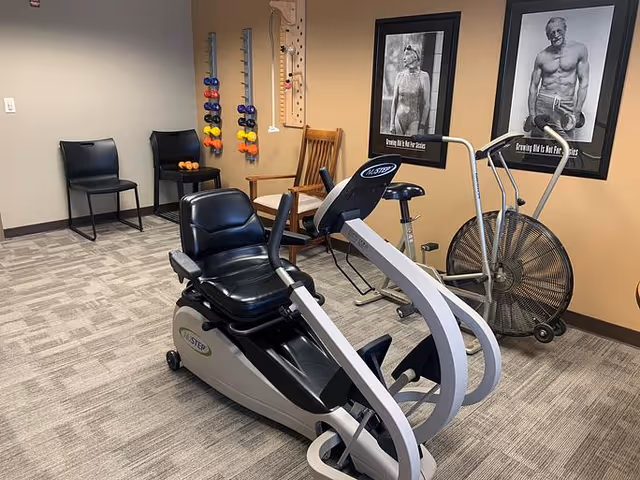 A small exercise room with a recumbent exercise bike, a stationary fan bike, and a wall-mounted rack holding colorful dumbbells. There are two black chairs and a wooden rocking chair against the wall. Two black and white motivational fitness posters featuring elderly individuals are hung on the wall.