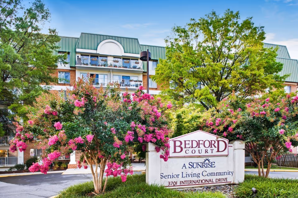 Exterior view of Bedford Court, a senior living community, showing a multi-story building with balconies and a green roof. In front of the building, there are blooming pink flowering trees and a sign that reads 'Bedford Court, A Sunrise Senior Living Community, 3701 International Drive.' The sky is clear and blue.