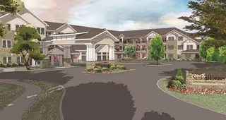 Exterior view of Sanford Estates Gracious Retirement Living facility showing a large multi-story building with a covered entrance, surrounded by trees and landscaped greenery under a partly cloudy sky.