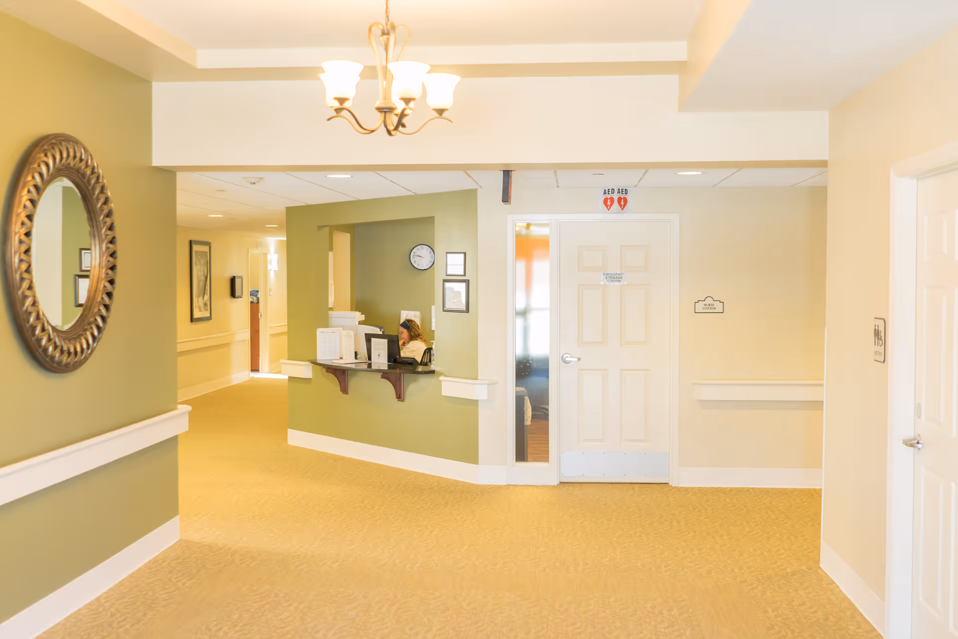 A bright hallway in a senior living facility with light green and beige walls, beige carpet, a decorative round mirror on the left wall, and a reception window where a staff member is seated at a computer. There is a white door with an AED sign above it and a small sign on the wall next to it. A chandelier with five lights hangs from the ceiling.