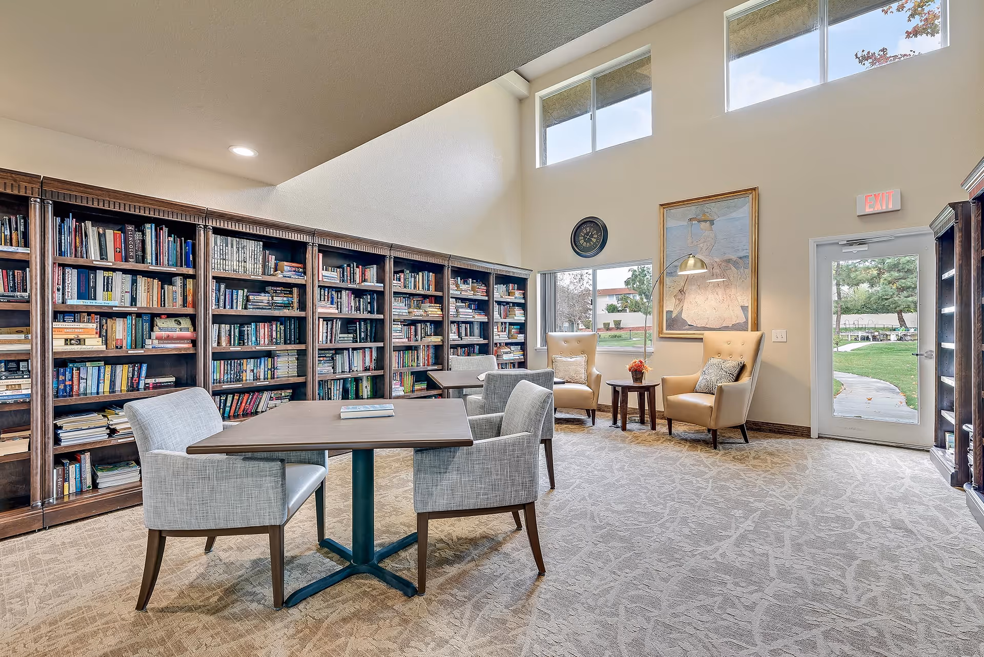 A bright and spacious senior living facility library with tall bookshelves filled with books along one wall. There are two tables with comfortable chairs for reading or socializing. Two beige armchairs with pillows are placed near a window and a door leading outside. The room has high ceilings with windows near the top, a wall clock, and a framed painting. The carpet has a subtle pattern, and natural light fills the space.