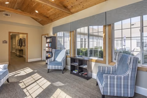 Sunlit sitting area with striped armchairs, a bookshelf and low shelving in front of large windows beneath a vaulted wooden ceiling.
