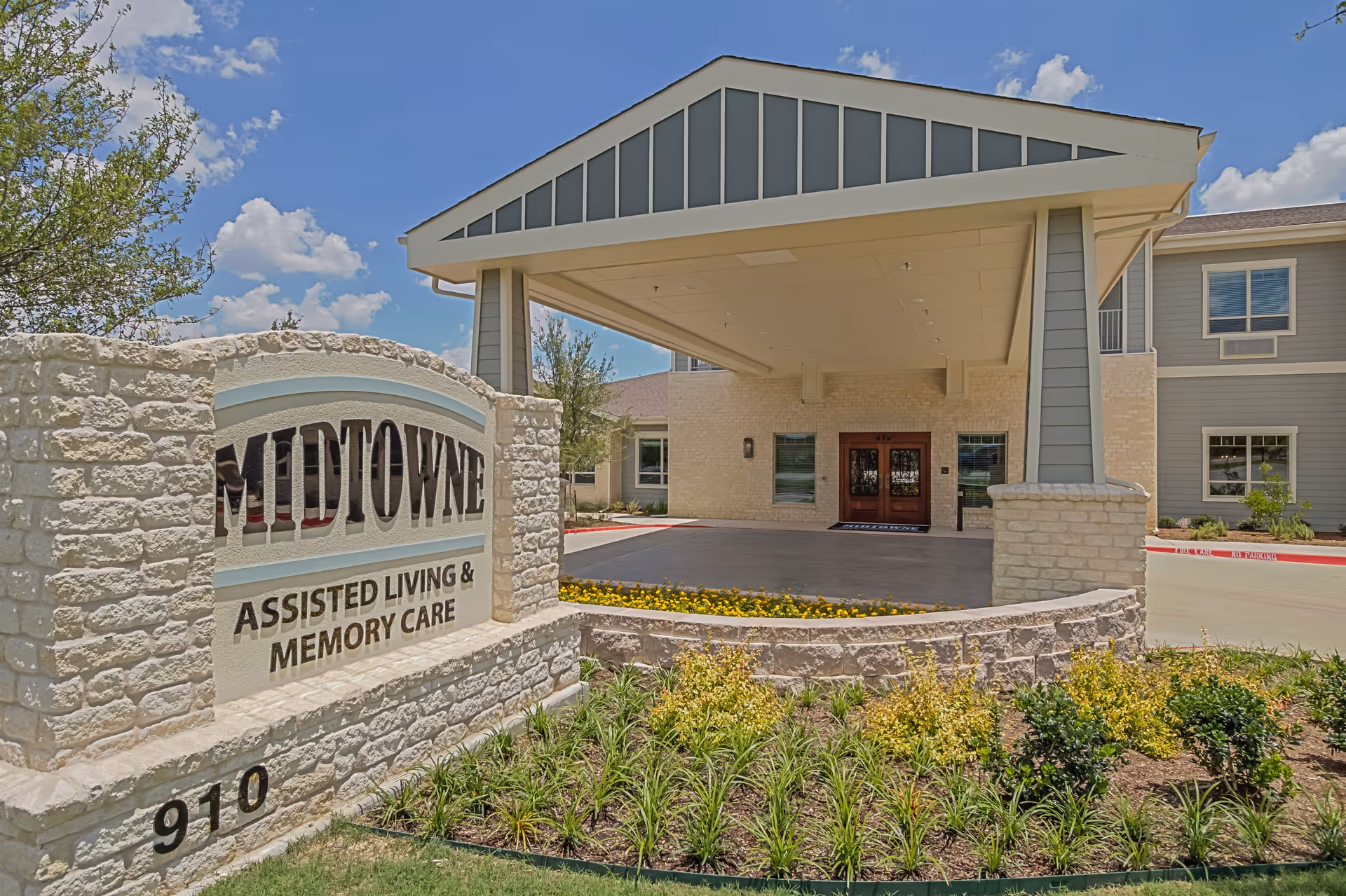 Exterior view of Midtowne Assisted Living and Memory Care facility showing the main entrance with a covered drop-off area, stone signage with the facility name, and landscaped greenery under a blue sky with some clouds.