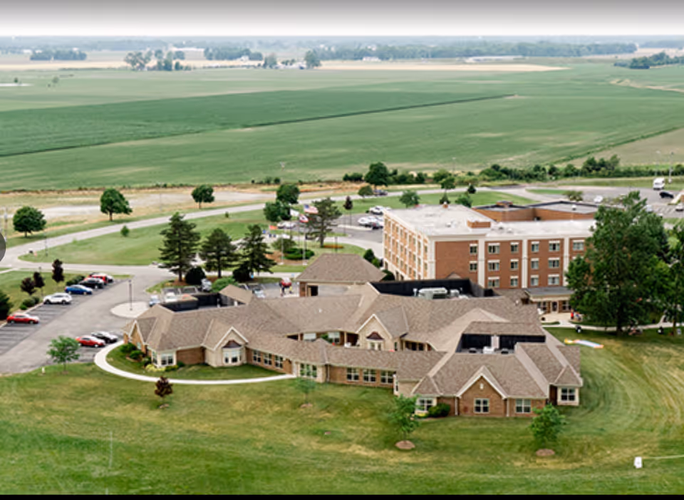 Aerial view of Browning Masonic Community showing multiple connected buildings surrounded by green lawns and trees, with a parking lot and open farmland in the background.
