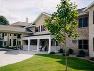 Exterior view of a two-story senior living facility building with beige siding and stone accents, a covered entrance with white pillars, a small tree and green lawn in the foreground under a partly cloudy sky.