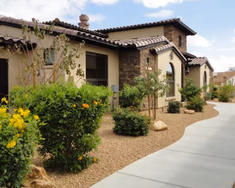 Exterior view of a single-story building with beige walls, stone accents, and a tiled roof. The building is surrounded by desert landscaping with bushes, small trees, and gravel. A curved concrete walkway runs alongside the building under a partly cloudy sky.