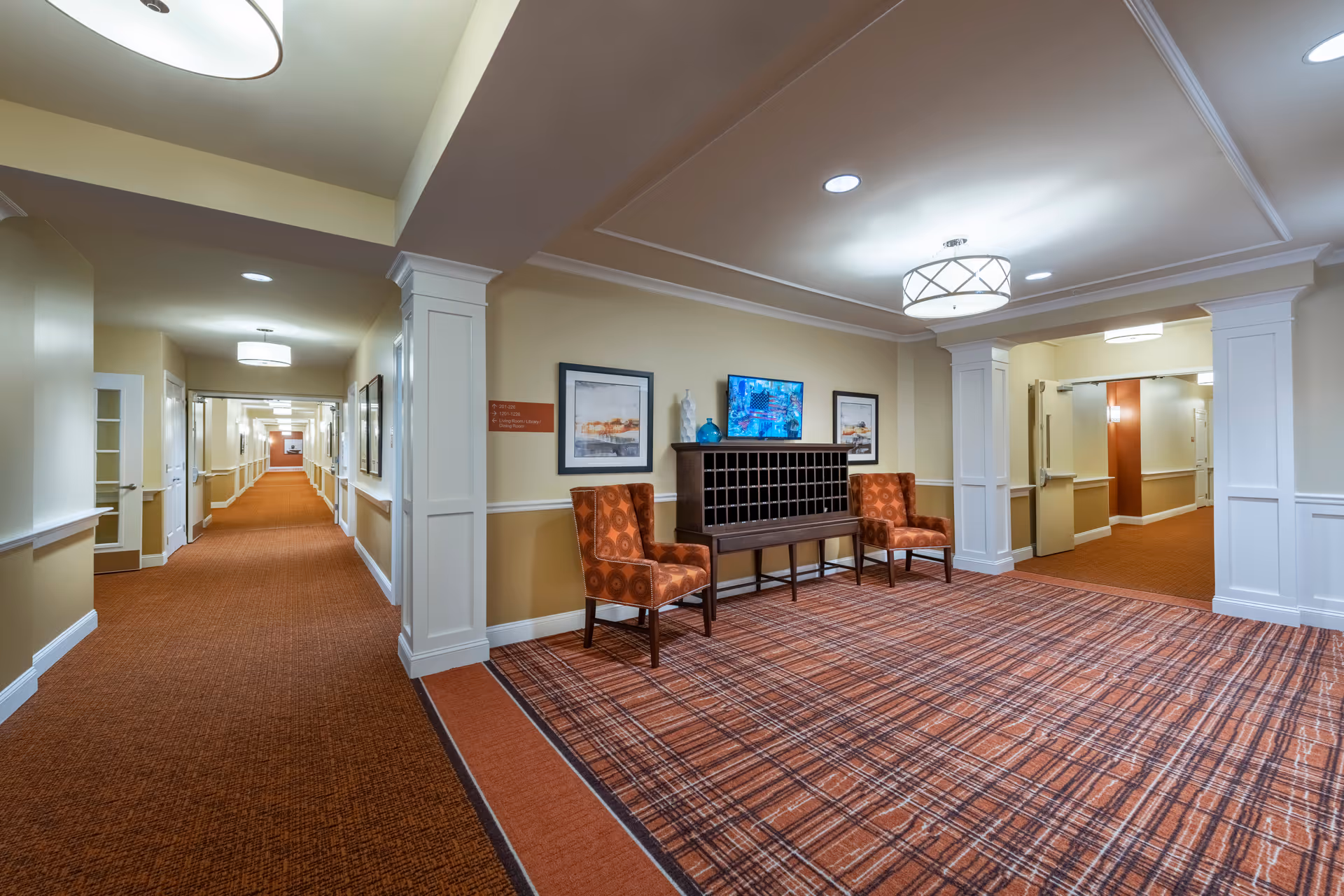 Interior hallway and seating area in a senior living facility with orange patterned carpet, two orange upholstered chairs, a wooden mail organizer, framed artwork on the walls, and ceiling lights.