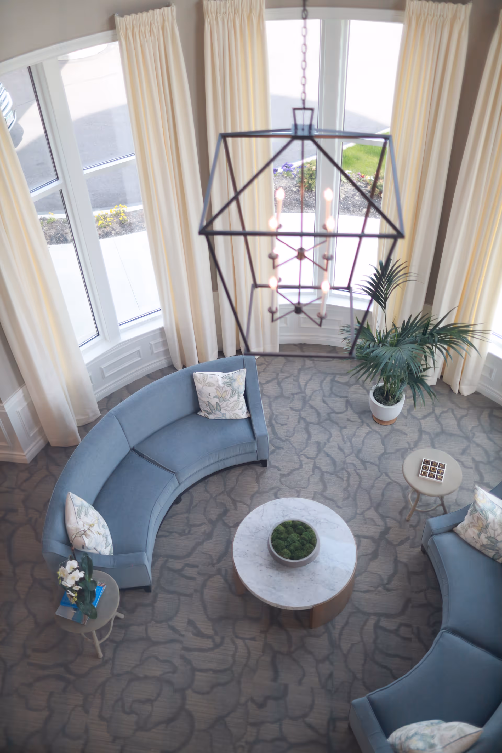 Overhead view of a living room area with two curved blue sofas, each with floral patterned pillows, a round marble coffee table with a bowl of green decorative moss, a small side table with a flower arrangement and books, a potted plant, large windows with cream-colored curtains, and a geometric chandelier hanging from the ceiling.