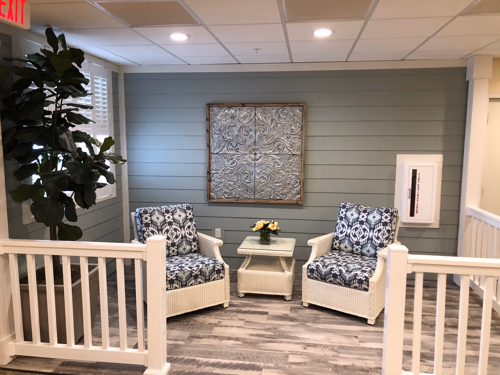 A cozy seating area with two white wicker armchairs featuring blue and white patterned cushions, a matching wicker side table with a small flower arrangement, a large potted plant to the left, and a decorative metal wall art piece on a light blue shiplap wall. The floor has a wood-like finish, and there is a white railing in the foreground.