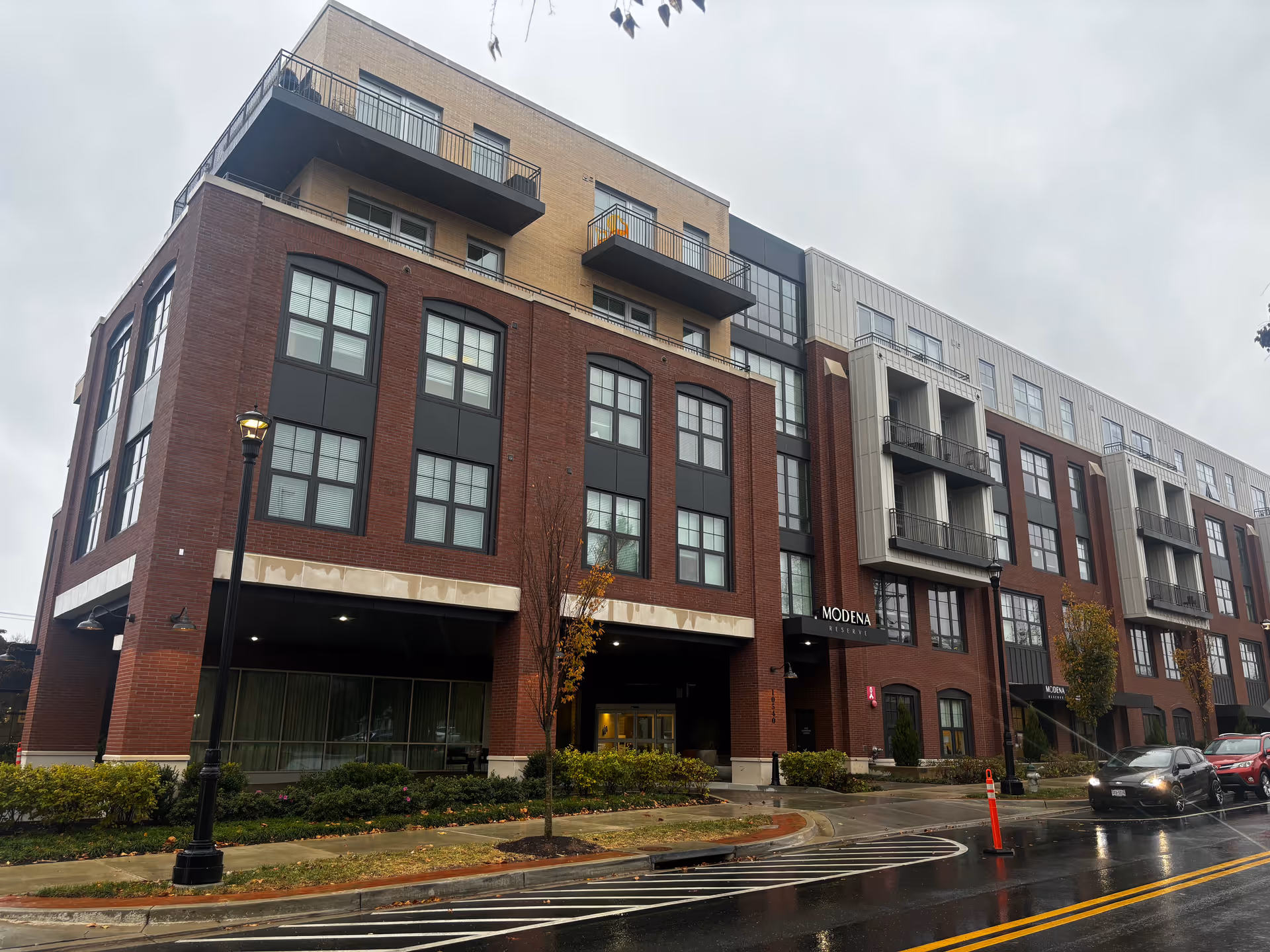 Exterior view of a multi-story brick residential building labeled "Modena" with balconies and a wet street in front on a rainy day.