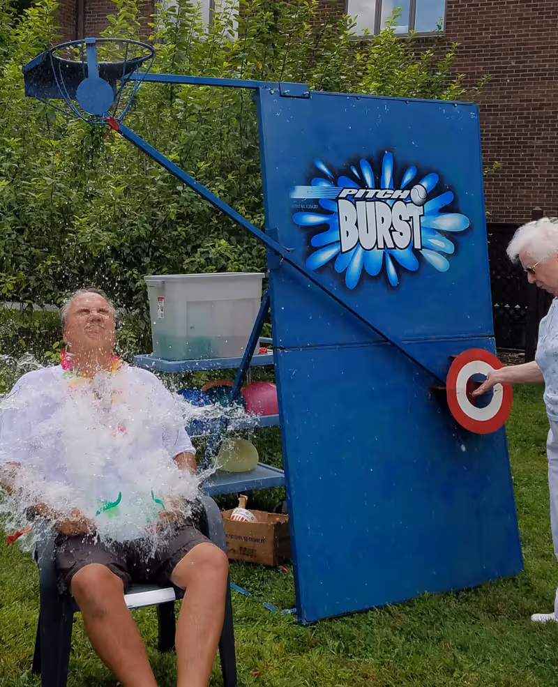 An elderly man sitting on a chair outdoors getting splashed with water from a dunk tank game called Pitch Burst, while an elderly woman stands nearby holding a target. The scene is set in a grassy area with trees and a brick building in the background.