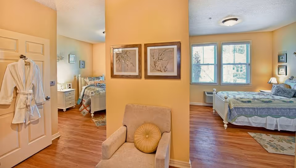 Interior of a senior living suite showing two bedrooms separated by a partial wall, a chair in the foreground, beds, nightstands, and a robe hanging on a door.