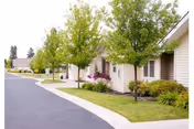 A row of single-story residential buildings with beige siding and brown roofs along a paved road. Small trees and well-maintained shrubs and grass line the sidewalk in front of the buildings under a clear sky.