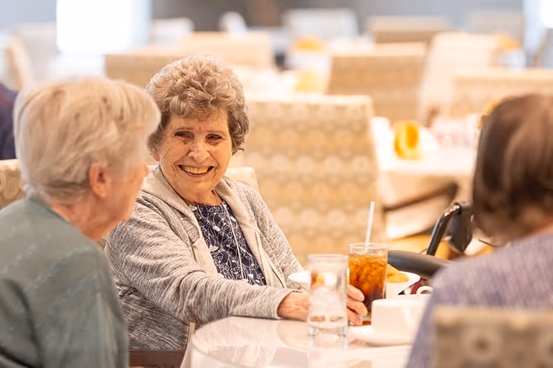 Three elderly women sitting and smiling at a table in a dining area, with drinks and plates in front of them. The background shows more tables and chairs with a warm, inviting atmosphere.