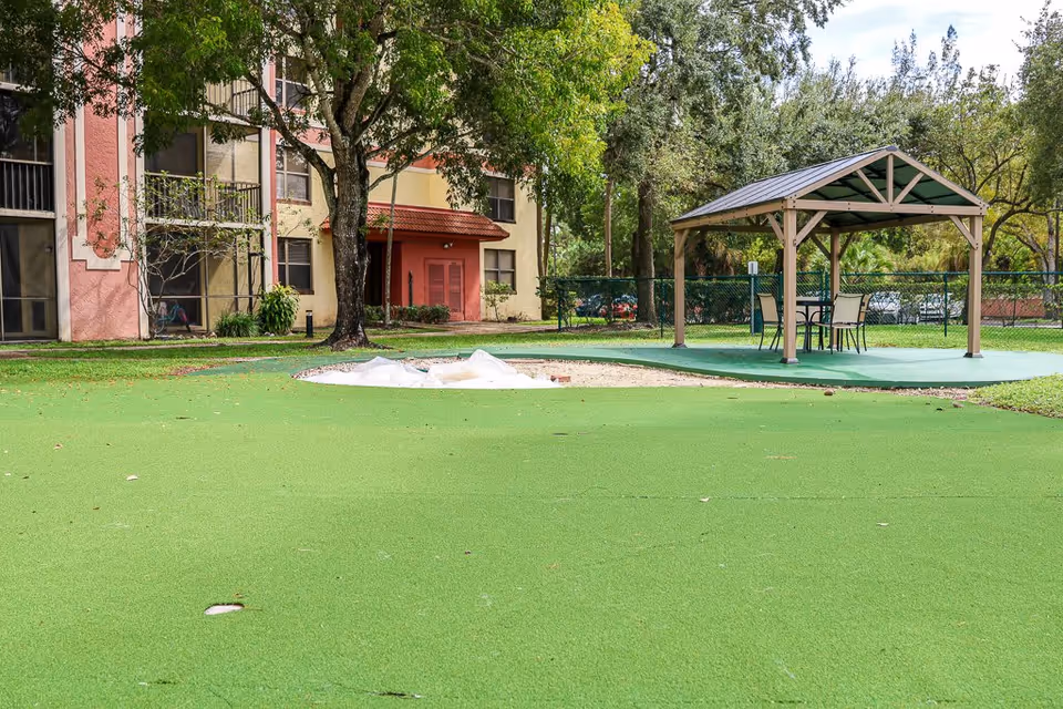 Outdoor area of a senior living facility featuring a green artificial turf surface with a small sandpit, a wooden gazebo with a metal roof, and a table with four chairs underneath. Trees and a multi-story building with balconies and windows are visible in the background.