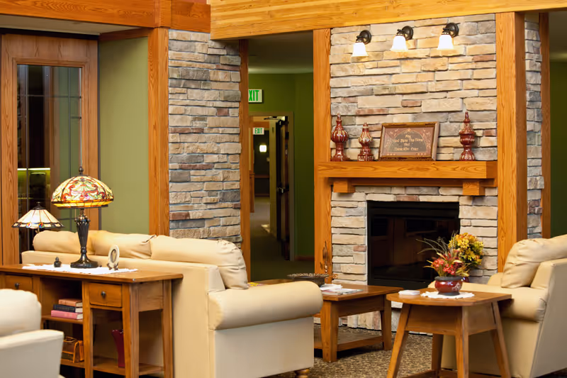 A cozy living room area with beige leather armchairs and wooden tables. There is a stone fireplace with a wooden mantle decorated with small vases and a framed sign. Two Tiffany-style lamps are on a wooden console table behind one of the armchairs. The walls are painted green with wooden trim, and there is a hallway with exit signs visible in the background.