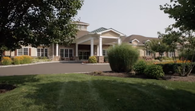 Senior living facility with a covered entrance, manicured lawn, and surrounding shrubs.