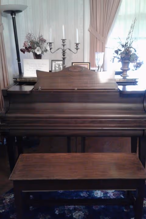 A wooden piano with a matching bench in front of it. On top of the piano are decorative items including a candelabra, framed photos, a glass, and floral arrangements. The background features light-colored curtains and a floor lamp.