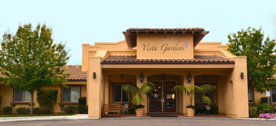 Front exterior view of Vista Gardens Memory Care facility with a beige stucco building, tiled roof, two potted palm plants flanking the entrance, a wooden bench on the left side, and trees on both sides of the building.