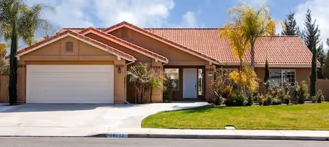 Single-story house with a red tiled roof, beige exterior walls, a two-car garage, and a well-maintained front yard with green grass, palm trees, and shrubs under a partly cloudy sky.